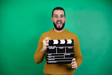 Handsome man wearing casual clothes over isolated green background holding clapperboard very happy...