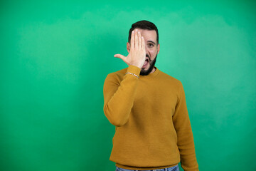 Handsome man wearing casual clothes over isolated green background covering one eye with hand,...