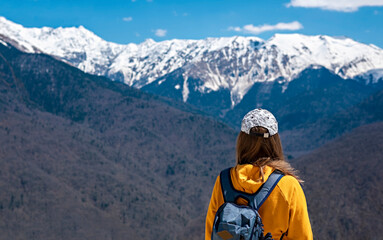 Rear view Young blond woman in yellow hoodie, cap, with backpack on background of snow capped Caucasus mountains. Hiking and travel, active lifestyle, copy space