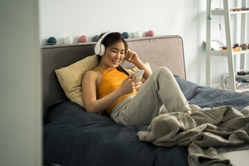 Woman wearing headphones laying at bed, listening music and looking at her smartphone