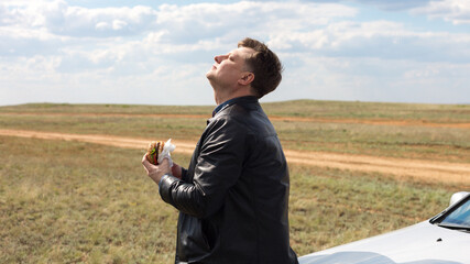 A man in nature in fresh air near the car eats a hamburger.