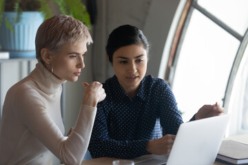 Pensive multiracial young businesswomen look at laptop screen brainstorm discuss company financial...