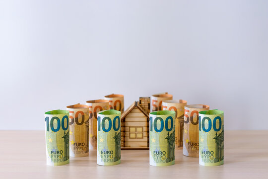 A small wooden house, surrounded on all sides by paper bills of 100 and 50 euros, rolled up one after the other. The concept of the budget of the real estate market. Investment in construction.
