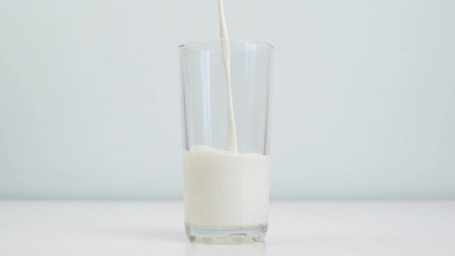 Milk is poured into a transparent glass. Cup of milk standing on the table on light background, close-up. Morning breakfast concept.