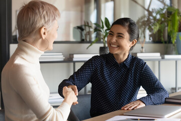 Overjoyed young Indian businesswoman shake hand of female colleague get acquainted greeting at...
