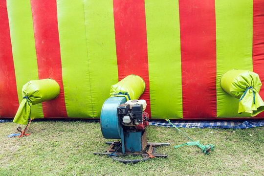 The old portable air blower pump fan install with green and red inflatable bouncer ball house castle or toy. Outdoor blue electric blower in the playground. Motor Air Blower on lawn in kid play land.