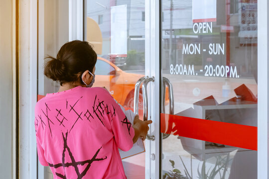 Back View Of Asian Woman Wearing Protective Face Mask To Prevent The Spread Of The Coronavirus (Covid-19) Holds A Parcel Box And Opens A Post Office Door To Deliver The Parcel. New Normal Lifestyle.