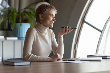 Smiling young woman worker sit at desk in office record audio voice message on modern smartphone. Happy successful Caucasian businesswoman talk on loudspeaker on cellphone. Communication concept.