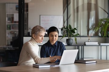 Young multiracial businesswomen sit at desk in office work together on laptop discuss ideas. Multiethnic diverse female employees colleagues brainstorm on computer at meeting. Teamwork concept.