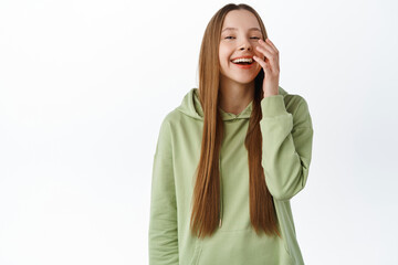 Positive candid girl with long hair and natural nude makeup, laughing, touching face and smiling happy, standing in hoodie against white background