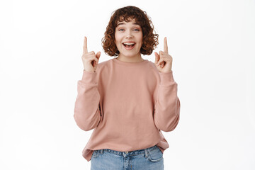 Happy and excited young girl looks amused, smiles at you while points up, shows cool advertisement,...