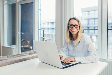 Portrait of a blonde woman looking at the camera sitting at home on the couch and holding a laptop