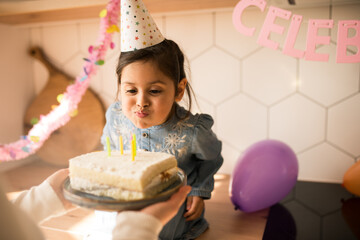 Girl wearing party cap blowing on the candles at her birthday cake