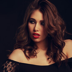 Fototapeta premium Beautiful bright makeup woman with long brown curly volume brown hair style, red lipstick, and black smokey eyes looking vamp and passion on black background. Closeup