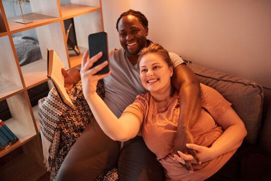 Man reading book and bonding to his lovely caucasian wife while she making selfie