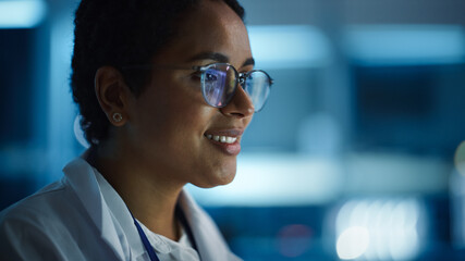 Beautiful Black Latin Woman Wearing Glasses Smiling Charmingly. Young Intelligent Female Scientist Working in Laboratory. Background Bokeh Blue with High-Tech Technological Lights