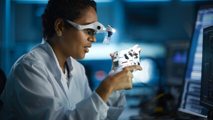 Modern Electronics Research, Development Facility: Black Female Engineer Wearing High-Tech Glasses Inspects Printed Circuit Board Motherboard. Scientist Designs Silicon Microchips, Semiconductors