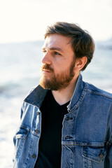 Portrait of a pensive man in a denim jacket looking into the distance against the background of the sea. Close-up