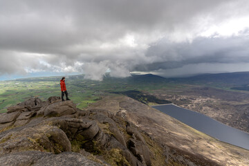 Man in red jacket standing on top of rocky mountain, from behind green fields, Irish Sea, spectacular clouds, Mourne Mountains North Ireland
