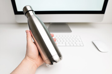 The male hand takes from the office desk steel eco thermo bottle for water. Metal water bottle. Silver color. Blank stainless steel double wall workout bottle. Empty stainless thermo water bottle. 