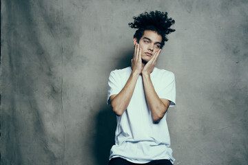 Cheerful guy with curly hair gestures with his hands near his face on a gray fabric background in a white t-shirt