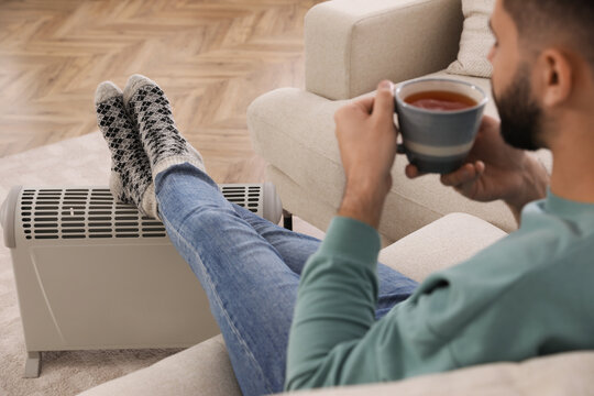 Young man with hot drink warming up near electric heater at home, focus on feet
