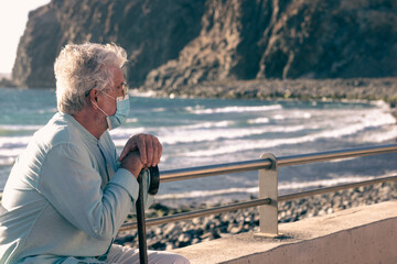 Fototapeta premium Coronavirus. Bearded old man sitting on the beach in the sunset light holding a walking stick because of body aches