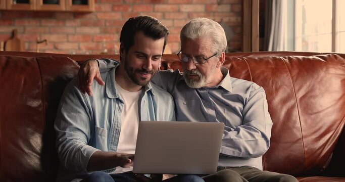 Older man his grown up millennial son sit on couch with laptop spend time together at home have fun on internet, younger gen teach older use modern digital tech. Buy online, e-commerce users concept