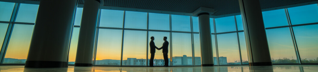 Fototapeta na wymiar The two men handshake in the office on panoramic window background