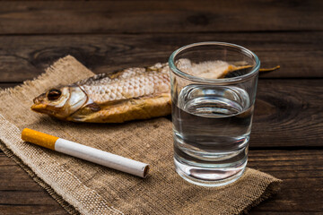 Shot of vodka with a stockfish and a piece of cloth with cigarette on an old wooden table. Close up view