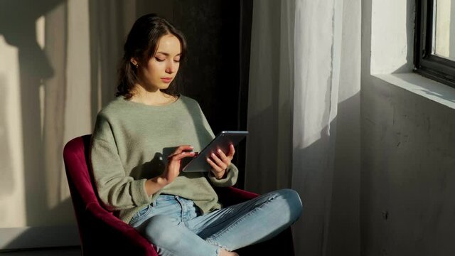 Beautiful woman sitting near window and using her tablet, reading phone message on the background of the sunrise.