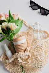 String bag with boquet of flowers, coffee, cup, glass bottle, hand sanitizer and sunglasses on white table. Summer fashion concept.