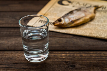 Shot of vodka with a stockfish and newspaper on an old wooden table. Close up view, focus on the shot of vodka