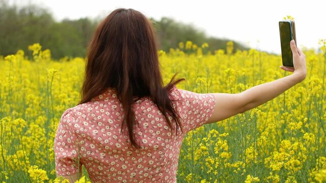 A beautiful cheerful young girl takes pictures of herself on the phone in a field against a background of yellow rapeseed flowers. A girl in a dress walks through a field of spring flowers.
