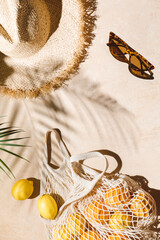 Summer flat lay on beige background. Straw hat, sunglasses, lemon fruits in eco friendly mesh shopping bag. Trendy palm shadow and sunlight, sun. Minimal summer travel fashion composition