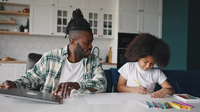 African american dad closing and putting away his laptop and talking to his drawing daughter, discussing picture at home