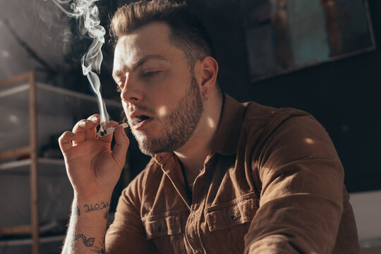 Man blowing out puffs of smoke while smoking weed hand rolled cigarette at home