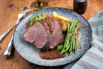 Traditional roasted venison roast with beens and potatoes in burgundy sauce served as close-up on a design plate - obrazy, fototapety, plakaty