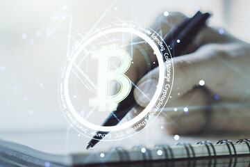Double exposure of creative Bitcoin symbol hologram with woman hand writing in notepad on background. Mining and blockchain concept