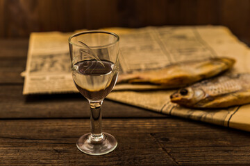 Glass of vodka with a stockfish and newspaper on an old wooden table. Angle view, focus on the glass of vodka