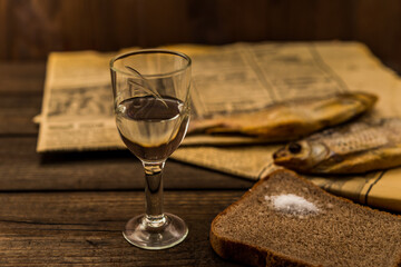 Glass of vodka with a stockfish and newspaper with piece of the black bread on an old wooden table. Angle view, shallow depth of field