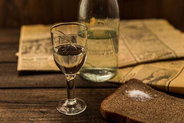 Glass of vodka with bottle and newspaper with piece of the black bread on an old wooden table. Angle view, shallow depth of field