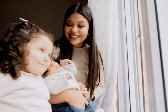 Young Hispanic mother with her 3-year-old daughter and her 1-month-old baby looking happily out the window. Mother and daughters at home