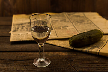 Glass of vodka with  newspaper and pickled cucumber on an old wooden table. Angle view, shallow depth of field, focus on the glass of vodka