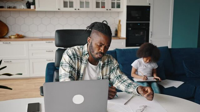 Parenthood and business. African american man working on laptop and writing notes, his little daughter drawing