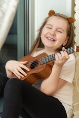 Portrait of happy smiling little girl in white t-shirt and black leggings sitting inside of home and playing ukulele. Hobbies and leisure activities concept. Vertical.
