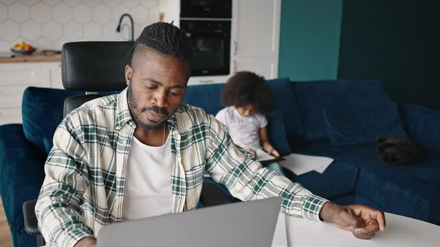 Close up of busy african american man working with laptop and documents at home office, his little daughter drawing