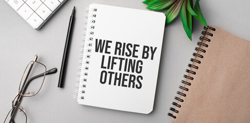 We Rise by Lifting Others is written in a white notebook with calculator, craft colored notepad, plant, black marker and glasses.