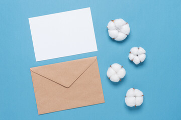 White blank card mockup with brown envelope and cotton flowers on paper blue background.