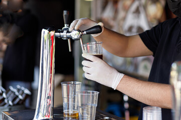Barman pouring fresh beer in plastic cup at street food festival, close up. Man hands in gloves pouring refreshing drink at counter in bar or pub. Summer vacation picnic. Street food market